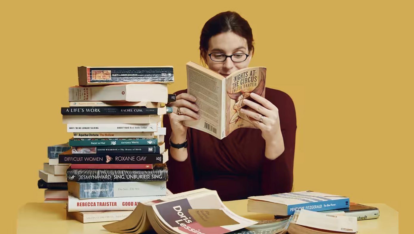 Woman reading a book with pleasure next to a stack of more books